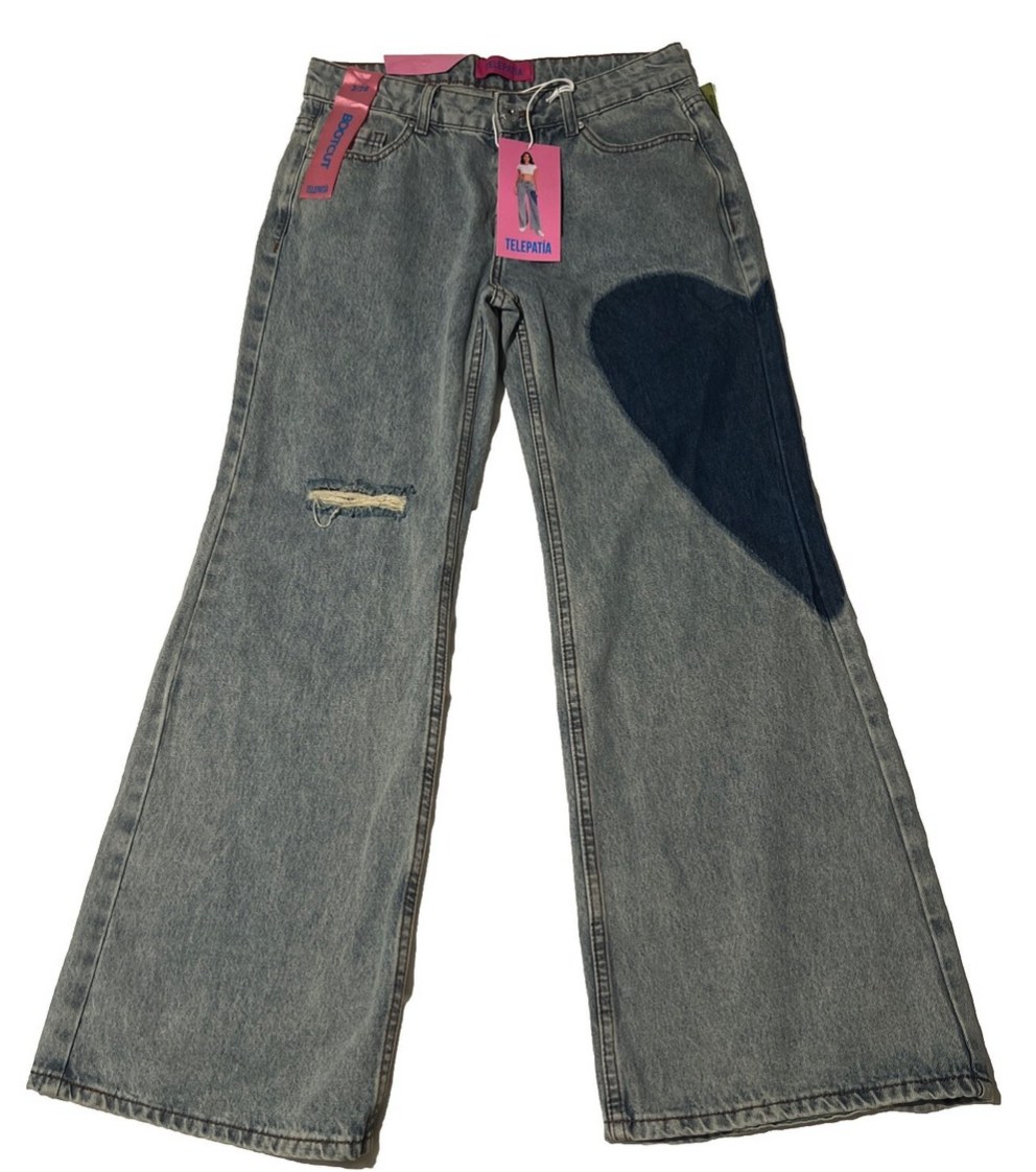 Telepatía Jeans grey bootcut jeans laid flat with a large dark denim heart patch on the right leg and a distressed knee rip, shown with pink brand hangtags