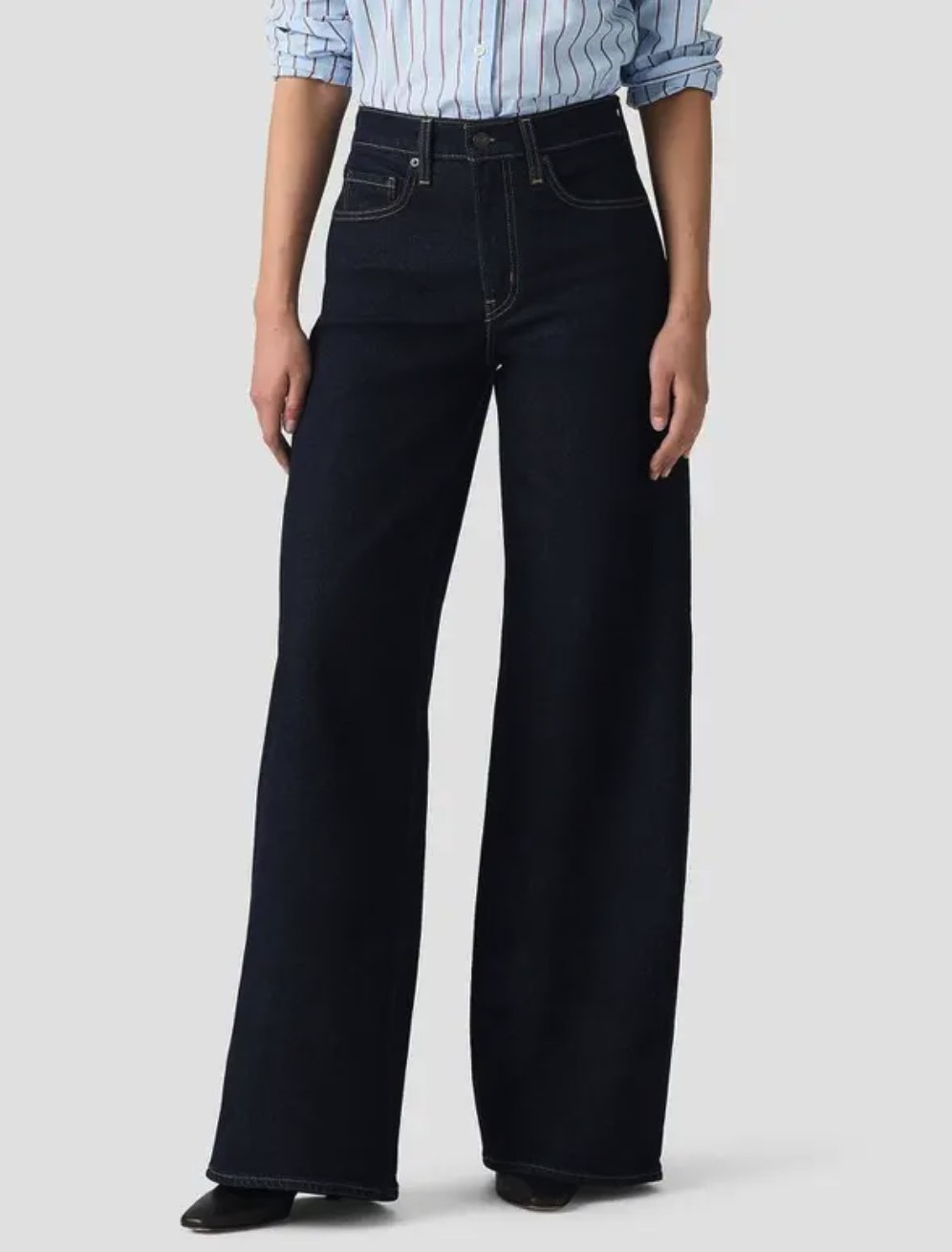 Woman wearing high-rise wide-leg dark indigo wash jeans with contrast stitching, paired with a light blue striped shirt and black shoes